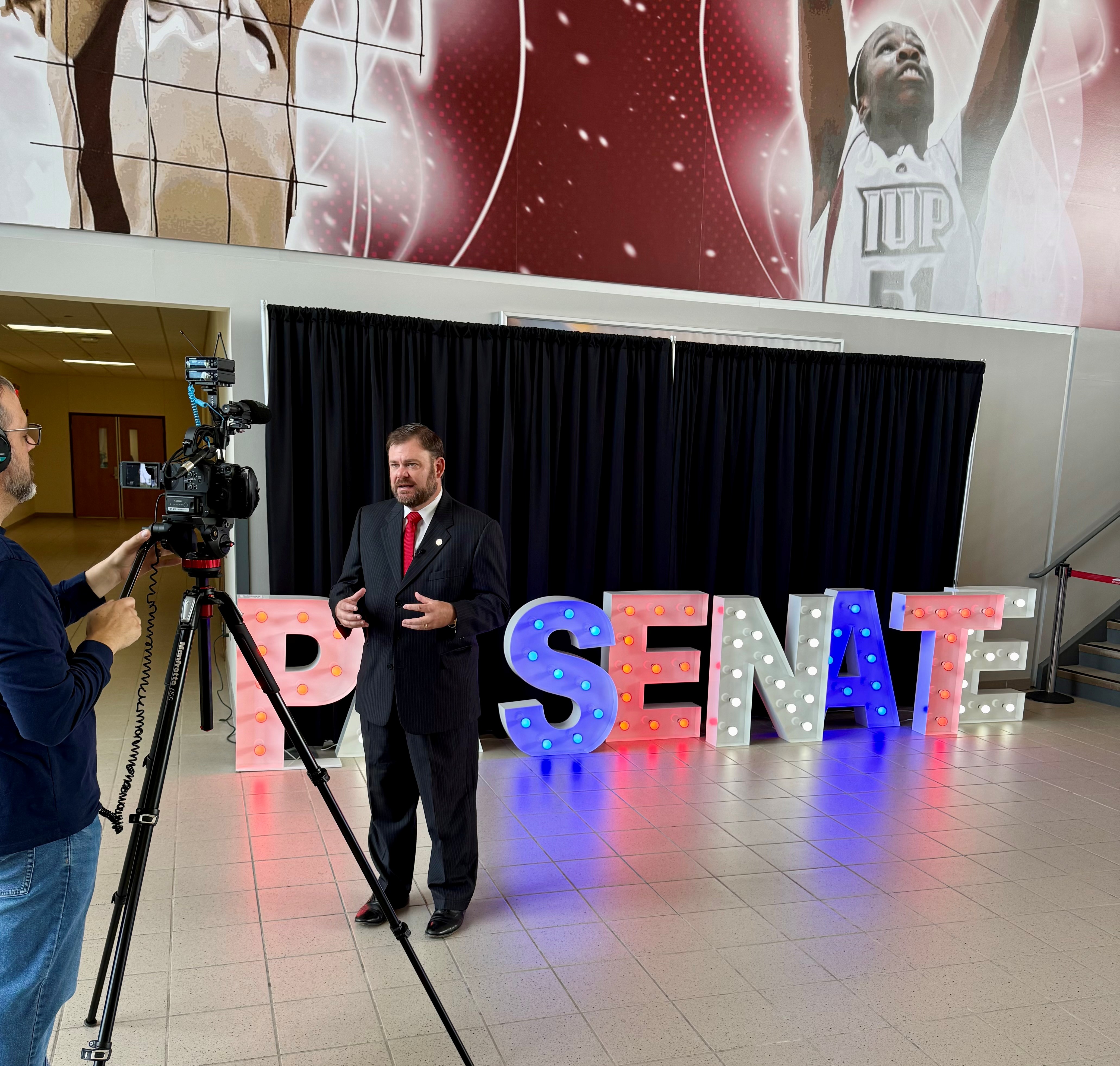 Sen. Pittman standing in front of large lighted letters spelling out PA Senate while being filmed by a camera man.